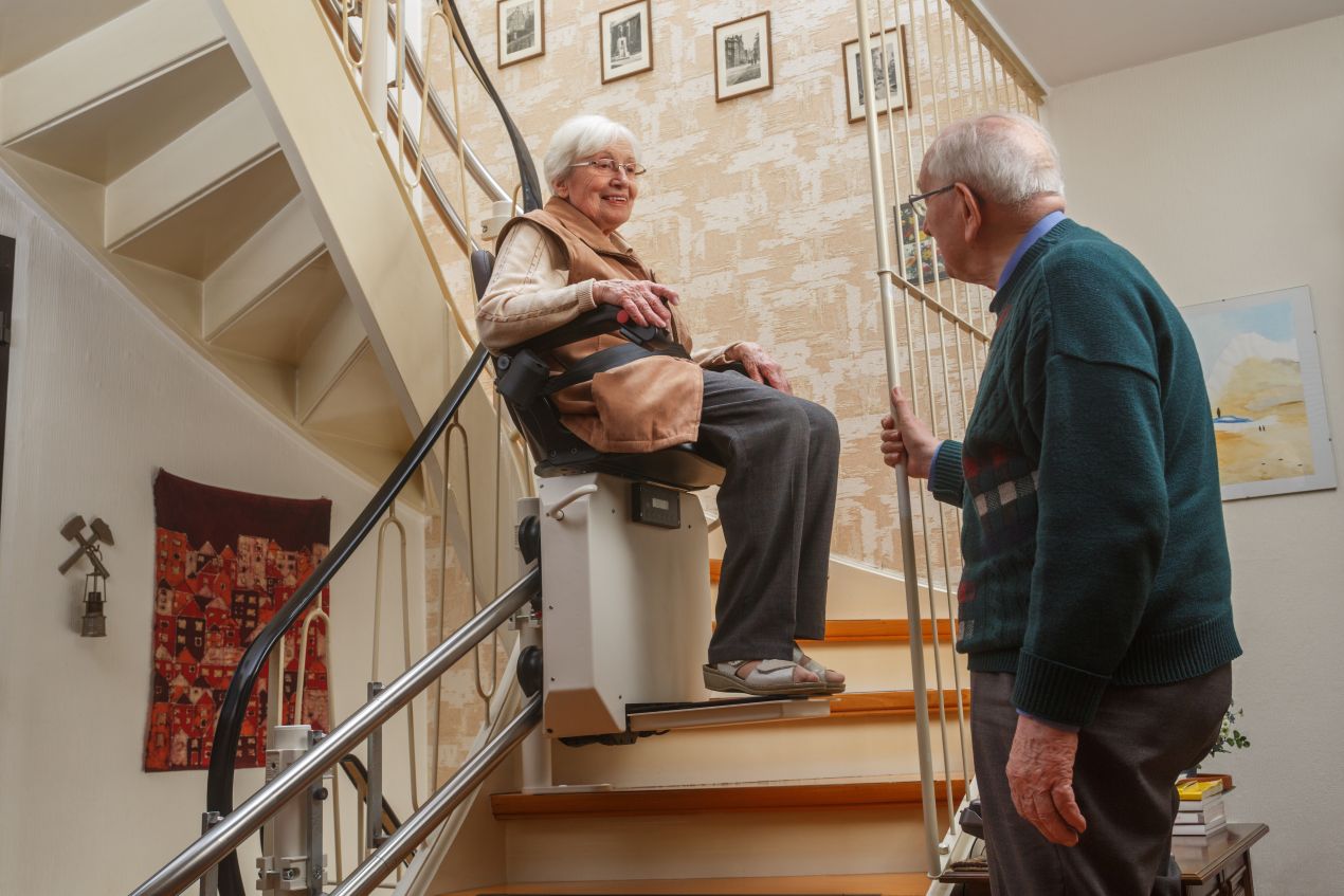 Ältere Frau nutzt Sitzlift an Treppe, während ihr Mann sie begleitet – barrierefreies Wohnen mit Sitzlift